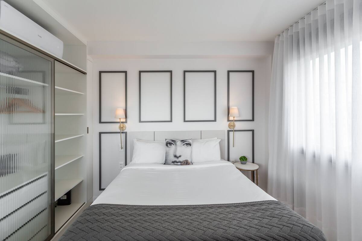 A modern hotel room with a large bed, a wardrobe, and a bedside table with a lamp.