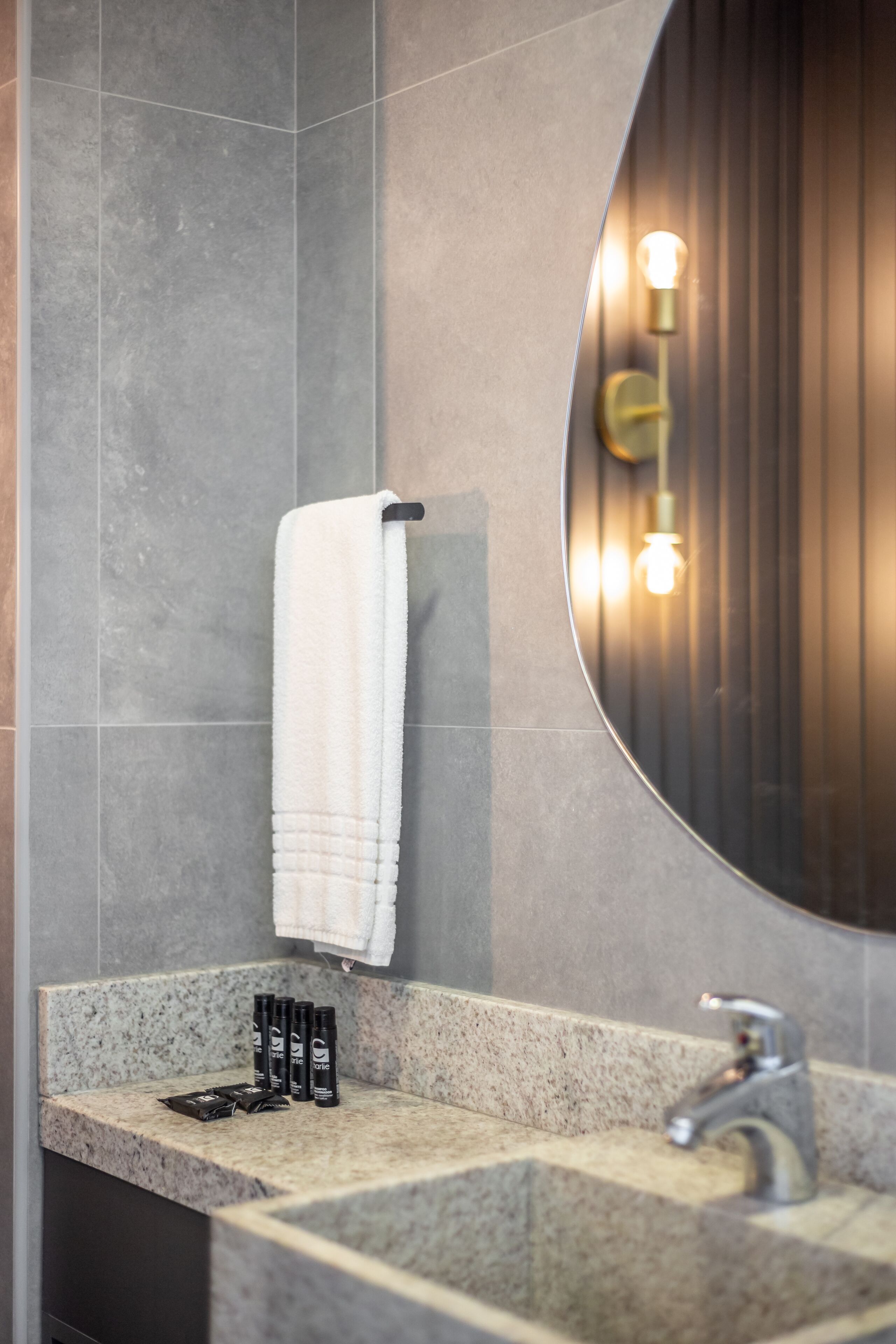 A bathroom with a granite countertop, a white towel hanging on a rack, a round mirror with a light fixture, and a faucet.