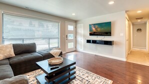 Una sala de estar con un sofá modular, una mesa de centro, un televisor montado en la pared y un ventanal con persianas.