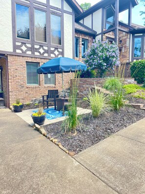 Uma casa residencial com área de pátio, mobiliário de exterior e um jardim com plantas e um guarda-sol azul.