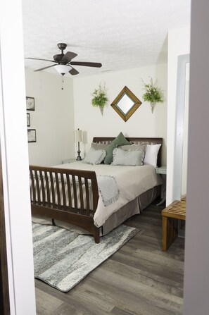 Quarto com cama de madeira, ventoinha no teto, mesa de cabeceira com abajur e duas plantas penduradas na parede.