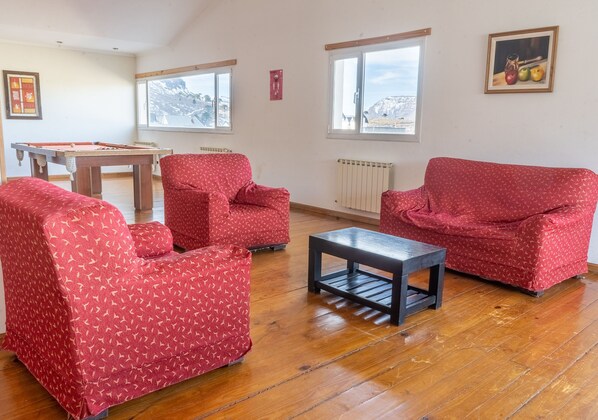 A room with a pool table, two red patterned armchairs, a small table, and a window with a view of a snowy landscape.