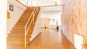 Una escalera de madera con barandilla de vidrio que sube a un segundo piso. En la izquierda, una pared blanca con un cuadro enmarcado y un letrero.