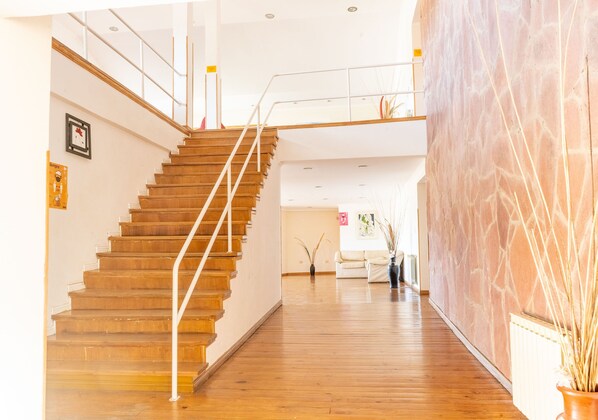 Una escalera de madera con barandilla de vidrio que sube a un segundo piso. En la izquierda, una pared blanca con un cuadro enmarcado y un letrero.