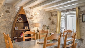 Ein rustikaler Essbereich mit einer Steinwand, Holzsesseln, einem Tisch mit gemustertem Tischläufer, einem Bücherregal aus Holz und einem Fenster mit Vorhängen.
