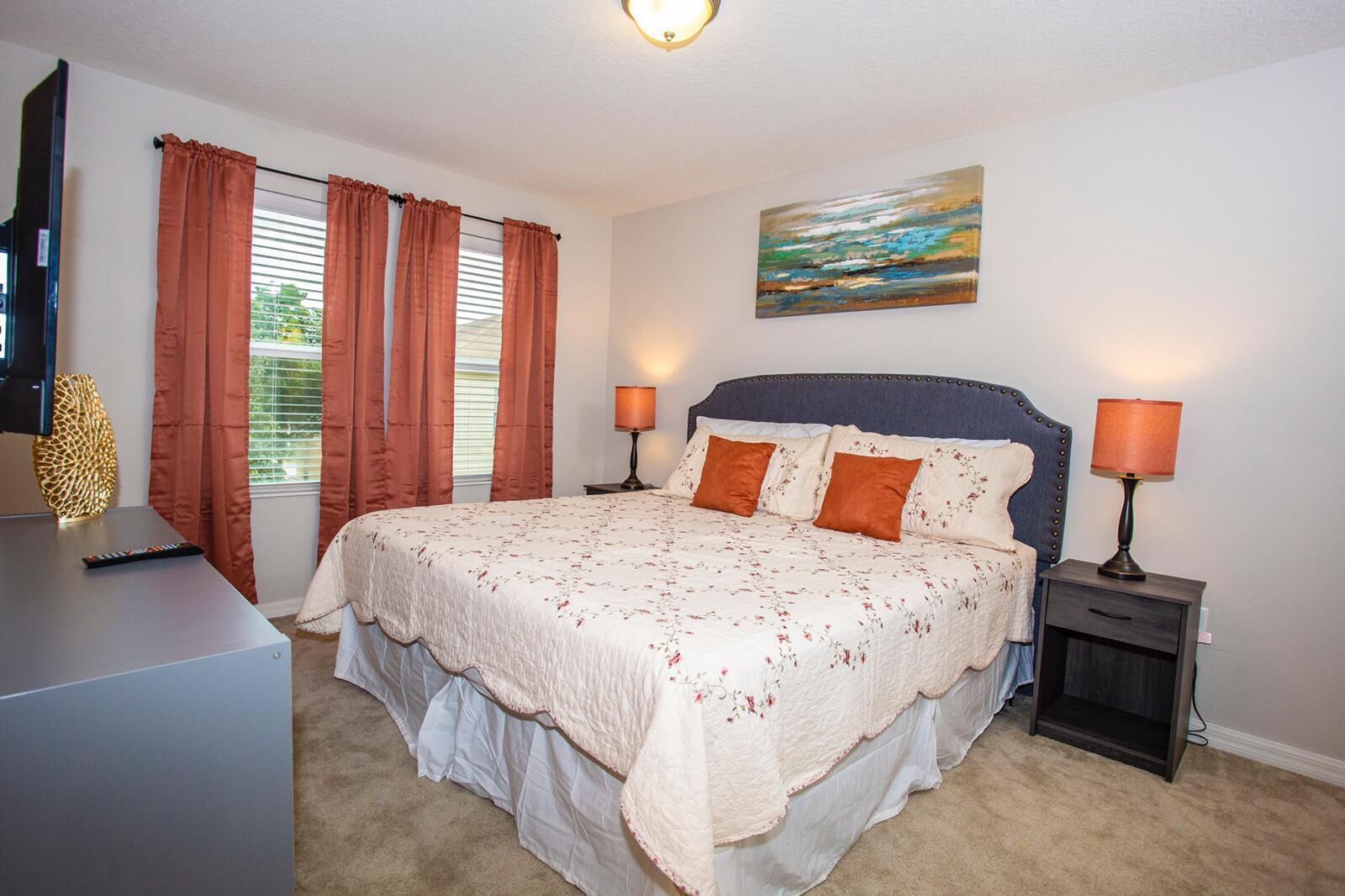A bedroom with a large bed, two bedside lamps, a painting on the wall, and a television on a stand.