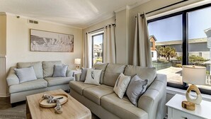 A living room with a sofa set, a coffee table, a side table, and a large window with curtains. - Oceanfront Balcony Views, Pool, STEPS to Beach Restaurants Shops (Wrightsville Beach)