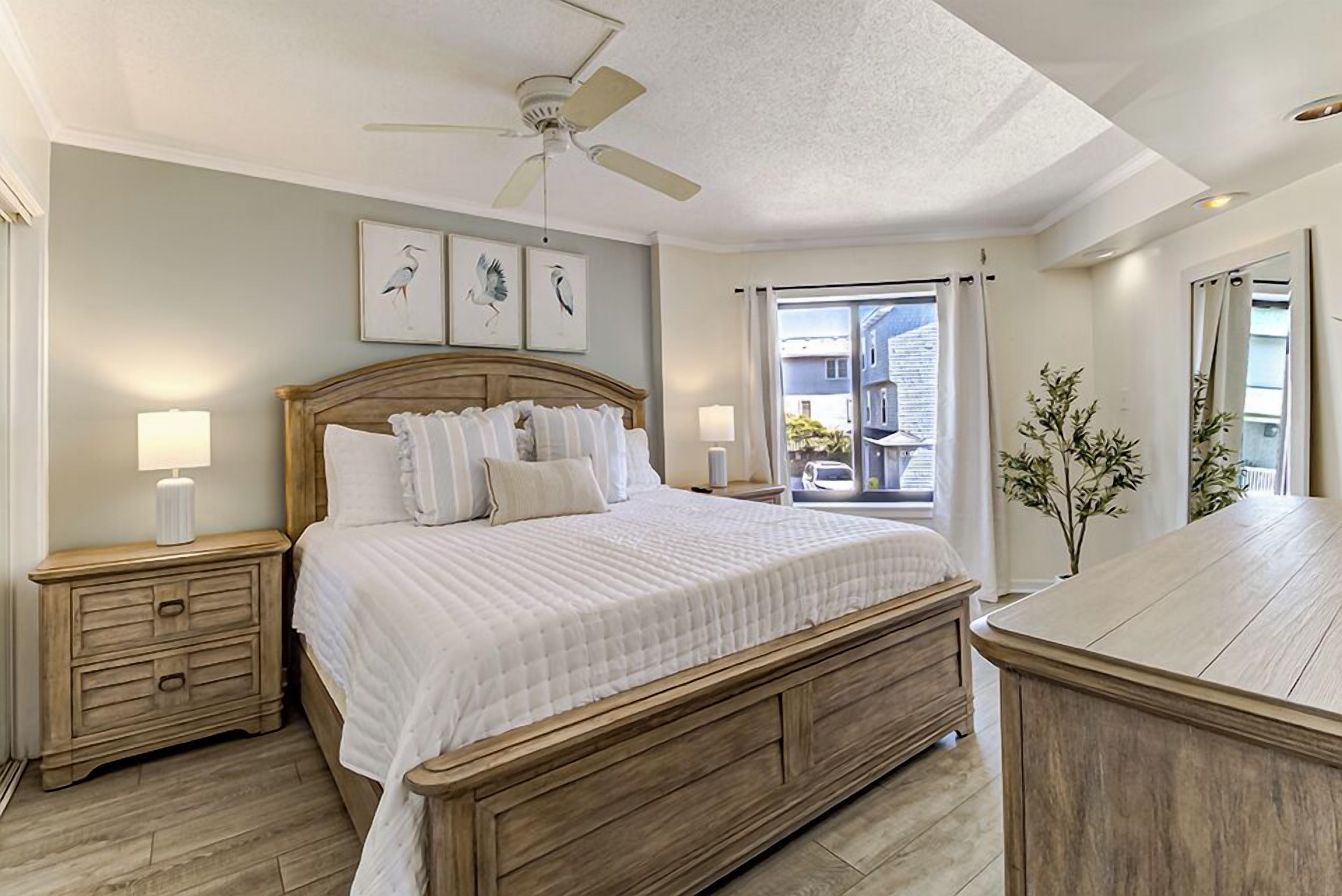 A bedroom with a wooden bed, two bedside tables with lamps, a ceiling fan, and a view of the outside.