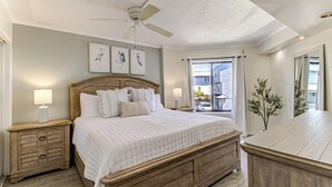 A bedroom with a wooden bed, two bedside tables with lamps, a ceiling fan, and a view of the outside. - Oceanfront Balcony Views, Pool, STEPS to Beach Restaurants Shops (Wrightsville Beach)