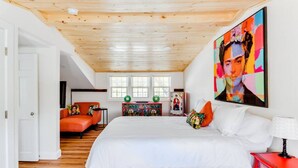 A bedroom with a wooden ceiling, a bed with white linens, an orange sofa, a colorful artwork on the wall, and a red nightstand.