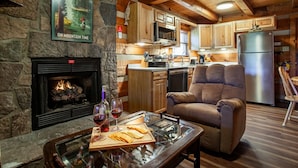 Interior acogedor de una cabaña con chimenea de piedra, armarios de madera, zona de cocina y área de estar confortable con una mesa de centro de vidrio.