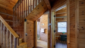 Interior de una cabaña de madera con una escalera que conduce a un segundo piso, un pequeño escritorio con una planta y una ventana con persianas.