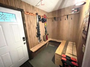 Interior de un refugio de esquí con paredes de madera, un banco, un soporte para esquíes y una mesa de madera con botas rojas.