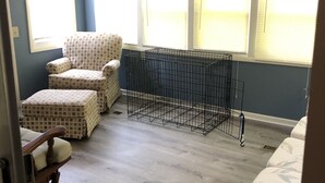 Una habitación con ventilador de techo, un sillón estampado, un pequeño reposapiés, una jaula para mascotas y una ventana con persianas.