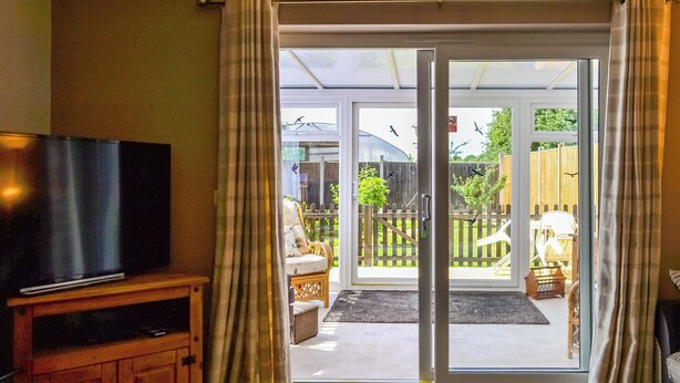 Una sala de estar con un televisor sobre un mueble de madera, un ventanal corredizo que da a un patio con mobiliario exterior y una cerca de madera.