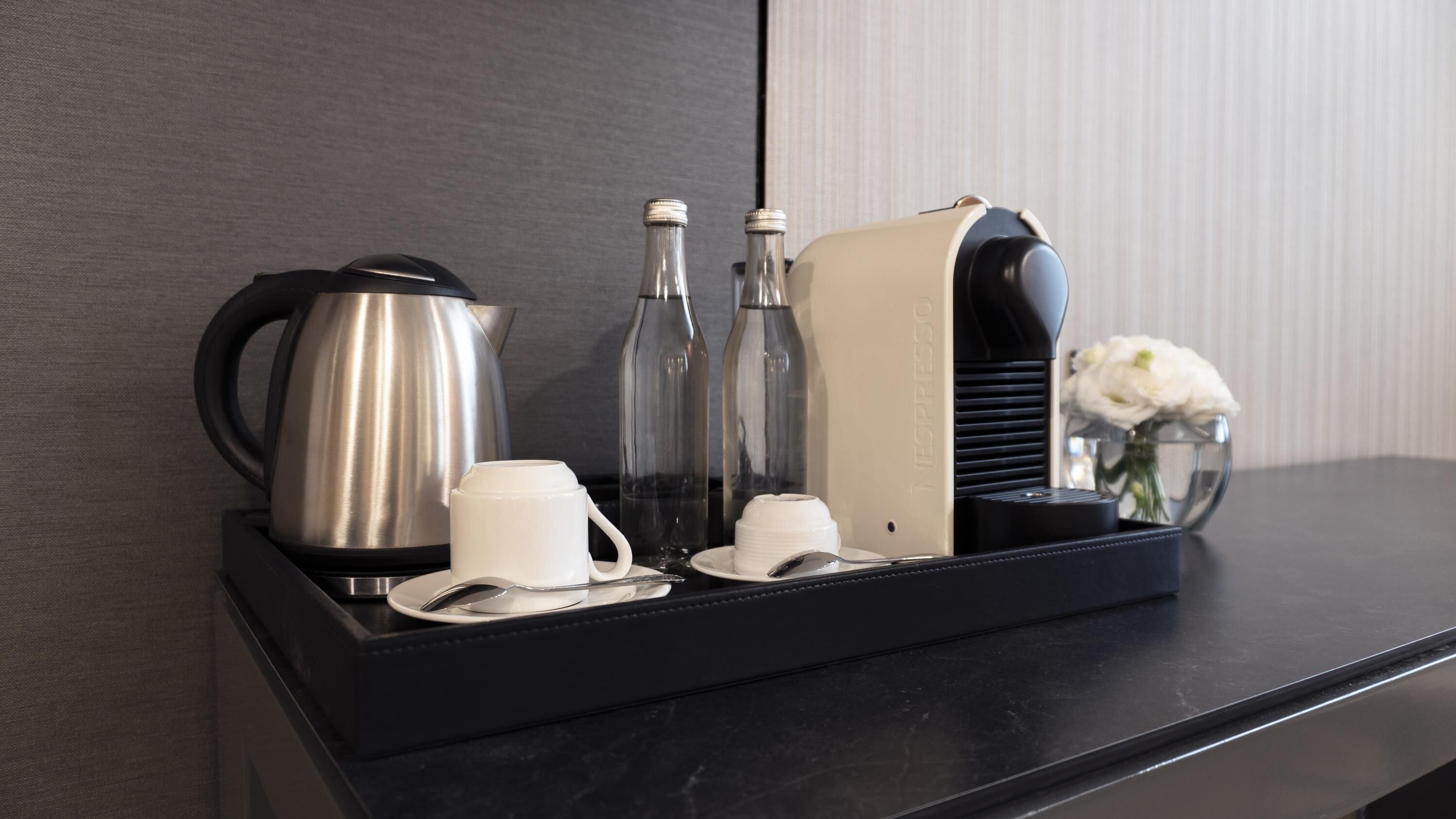 A tray with a coffee maker, two bottles, a cream dispenser, and a vase with flowers on a black countertop.