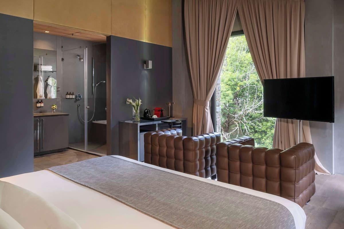 A modern hotel room with a large bed, a seating area with cushioned chairs, a small desk with a flat-screen TV, and a view of greenery outside.