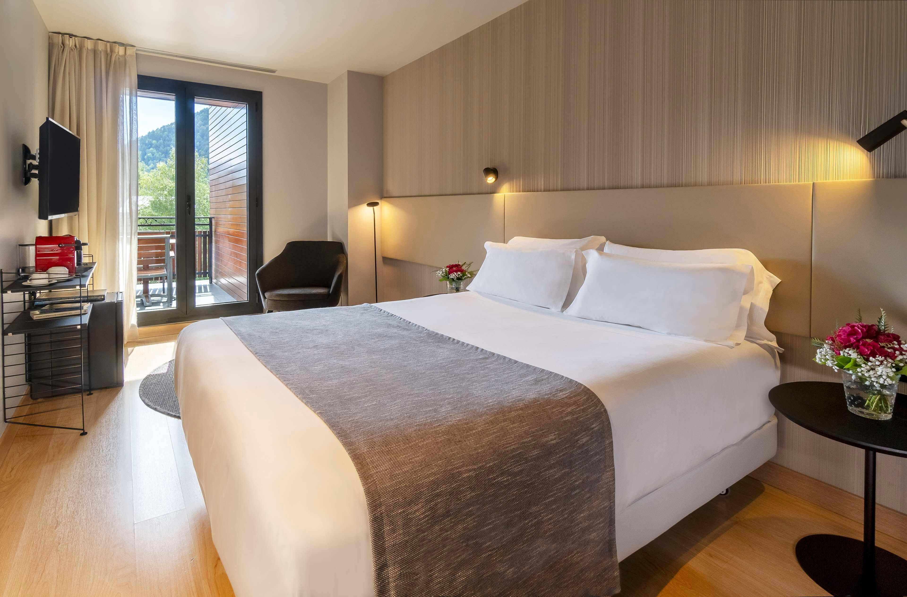 A hotel room with a large bed, a desk with a red object, a chair, a small table with flowers, and a balcony with a view.