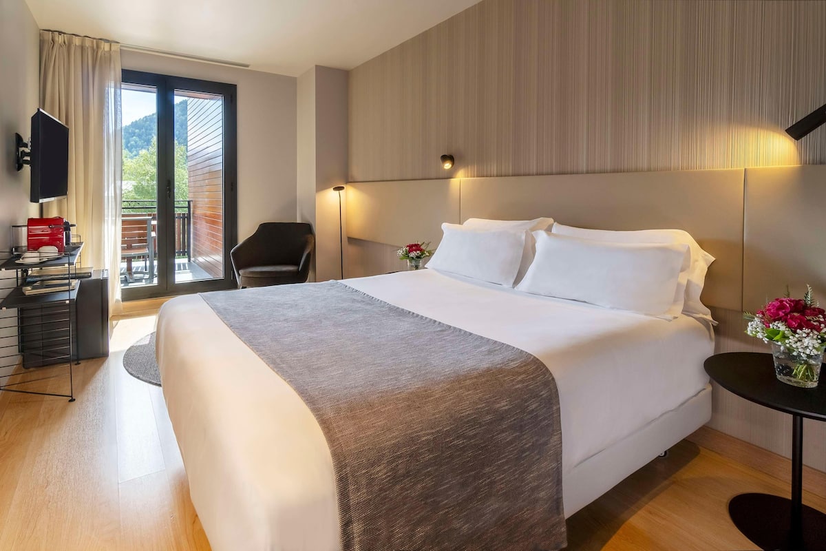 A hotel room with a large bed, a desk with a red object, a chair, a small table with flowers, and a balcony with a view.