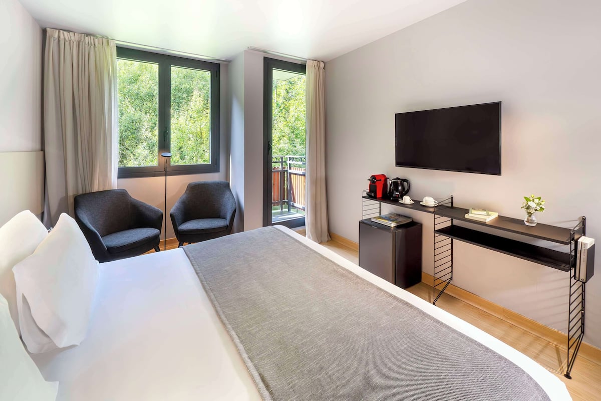 A modern hotel room with a large bed, two chairs, a desk with a flat-screen TV, and a balcony with a view of trees.
