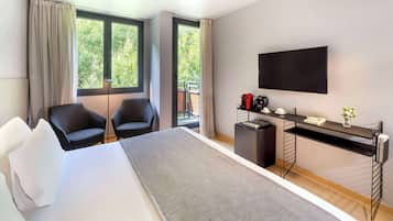 Une chambre d’hôtel moderne dotée d’un grand lit, de deux fauteuils, d’un bureau avec un téléviseur à écran plat et d’un balcon offrant une vue sur les arbres.