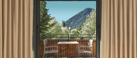 A room with a balcony featuring a wooden table and two chairs, a view of a mountain, and curtains on either side.