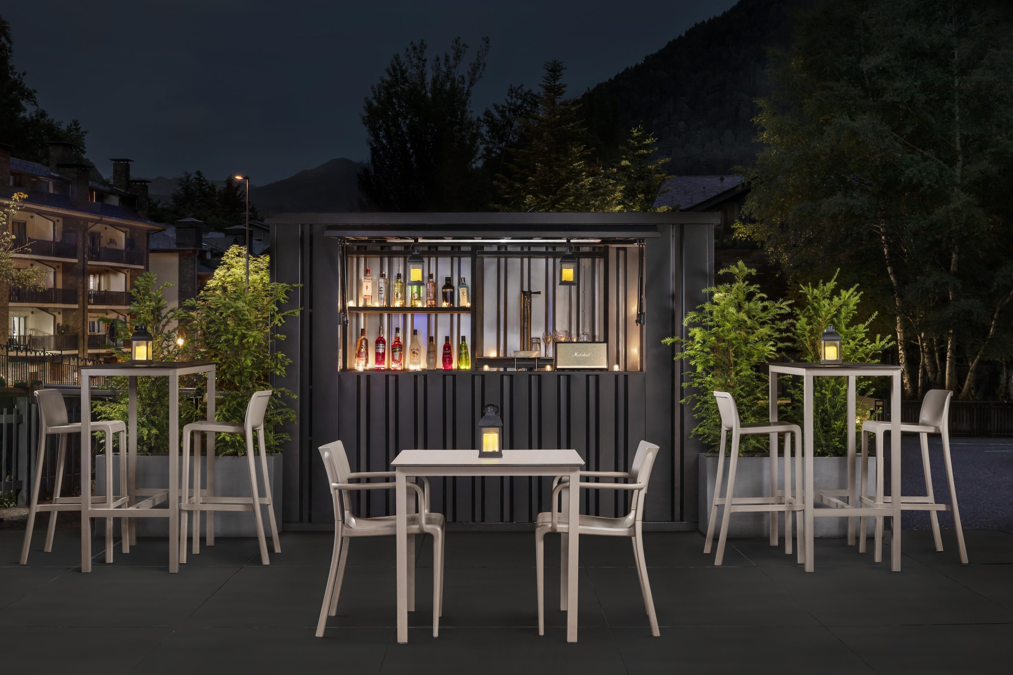 An outdoor seating area with a table and chairs, a bar with bottles, and a view of buildings and trees.