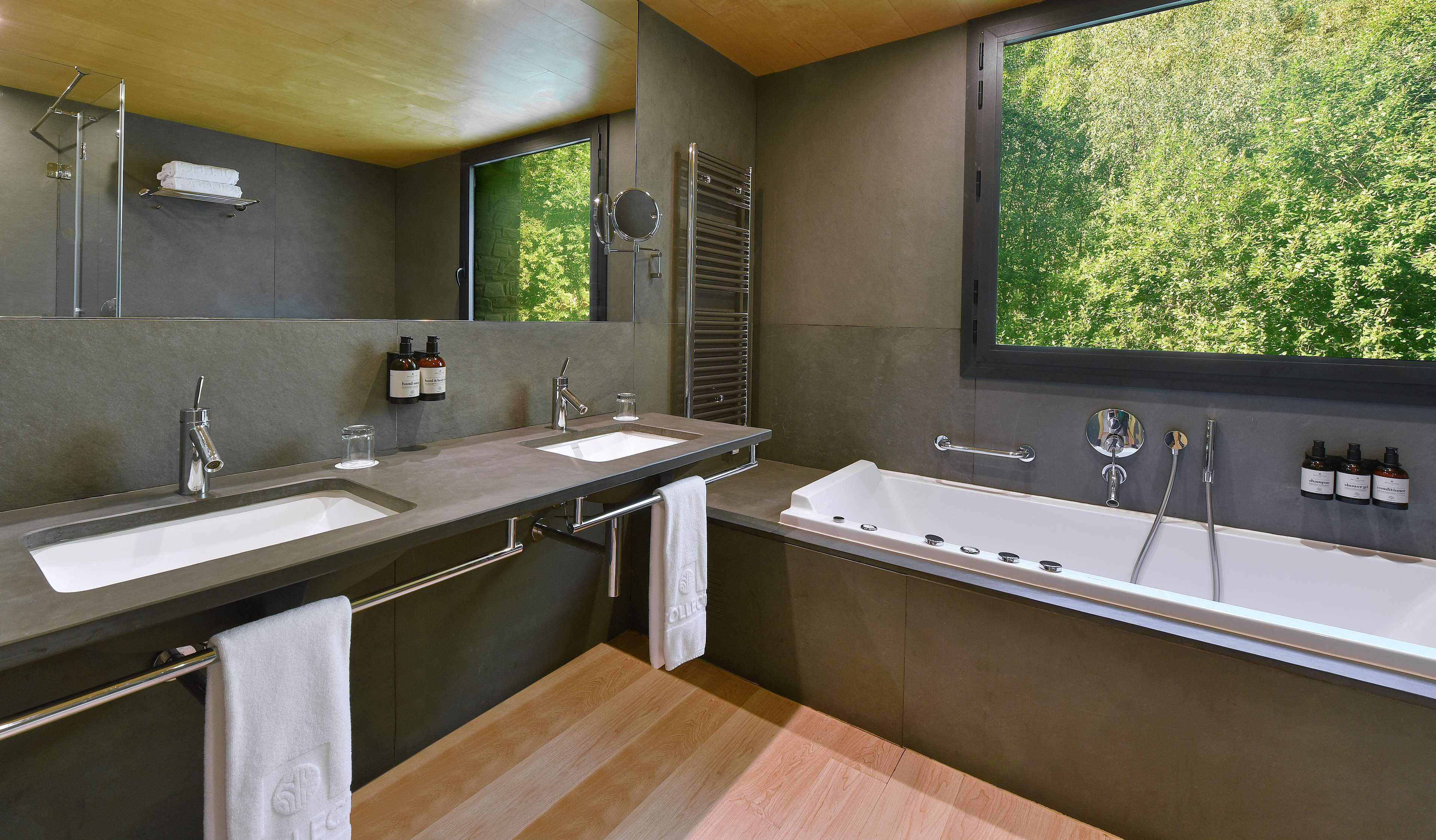 A modern bathroom with two sinks, a bathtub, and a large window with a view of greenery.