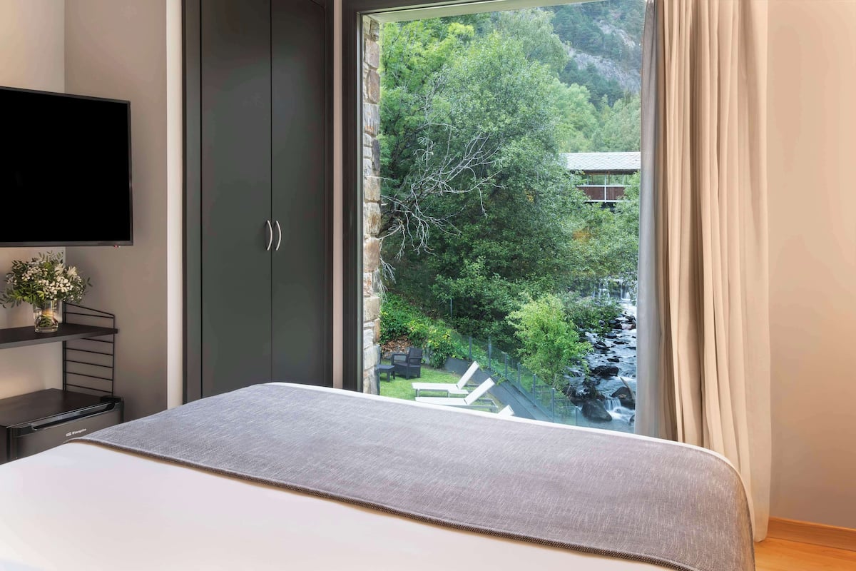 A bedroom with a bed, a TV, a wardrobe, and a window with a view of greenery and a building.