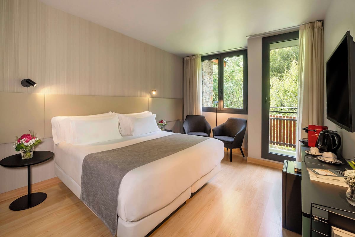A modern hotel room with a large bed, a small table with a vase of flowers, a chair, a desk with a TV, and a window with a view of greenery.