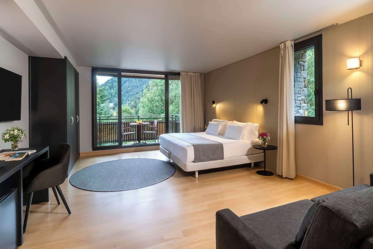 A modern hotel room with a bed, a desk, a chair, a small table, and a balcony with a view of the forest.