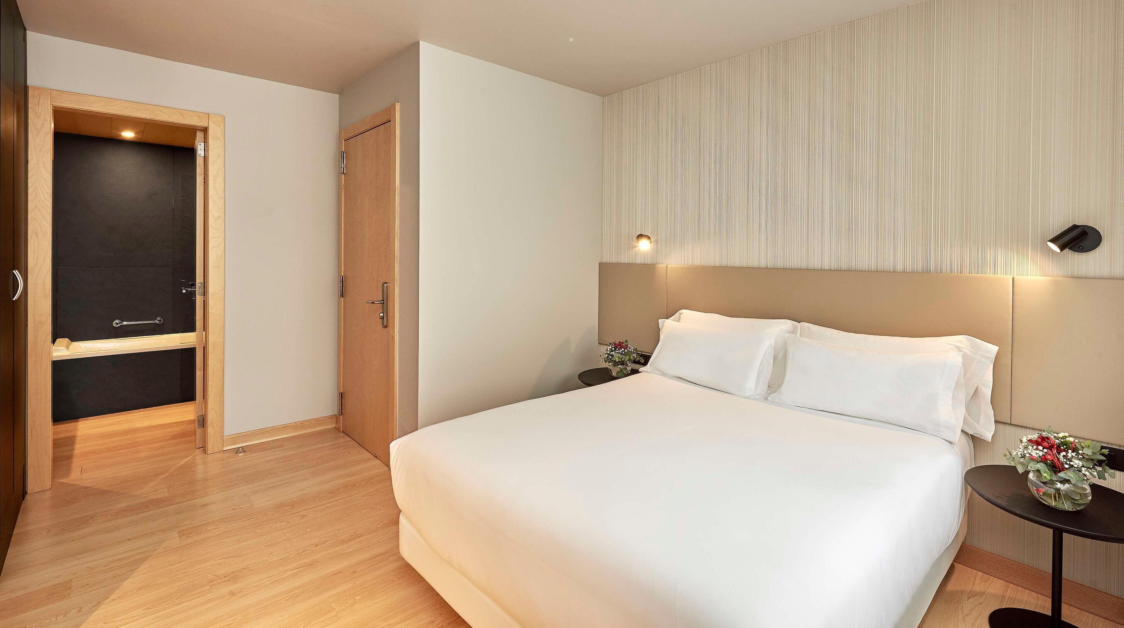 A hotel room with a bed, a wooden wardrobe, a small table with a vase, and a wall-mounted light fixture.