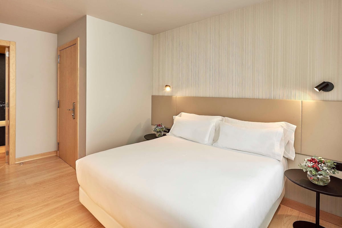 A hotel room with a bed, a wooden wardrobe, a small table with a vase, and a wall-mounted light fixture.