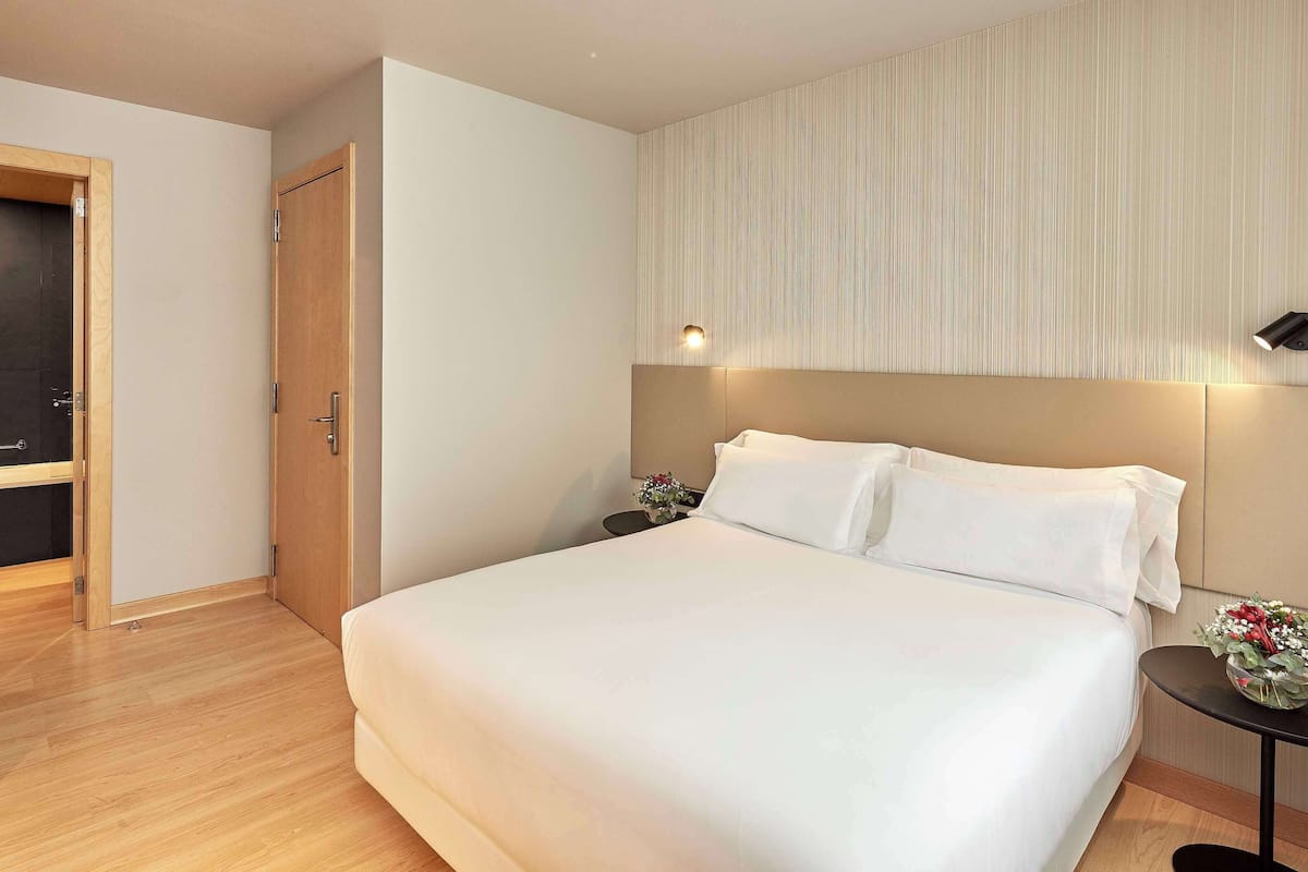 A hotel room with a bed, a wooden wardrobe, a nightstand, and a wall-mounted light fixture.
