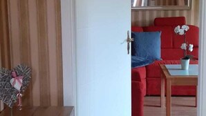 Een kamer met een houten bank, een witte deur en uitzicht op een zithoek met een rode bank, een blauw kussen en een kleine tafel met een plant.