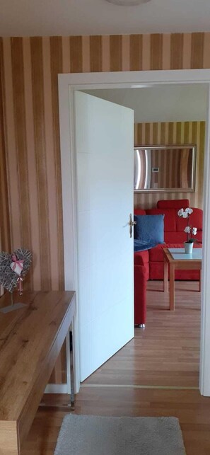 A room with a wooden bench, a white door, and a view into a living area with a red sofa, a blue pillow, and a small table with a plant. - Vacation bungalow Wiesenblick (Lambrechtshagen)