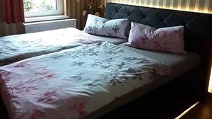 Een tweepersoonsbed met bloemenbeddengoed, een hoofdbord met ingebouwde verlichting, een raam met gordijnen en een gestreepte wand.