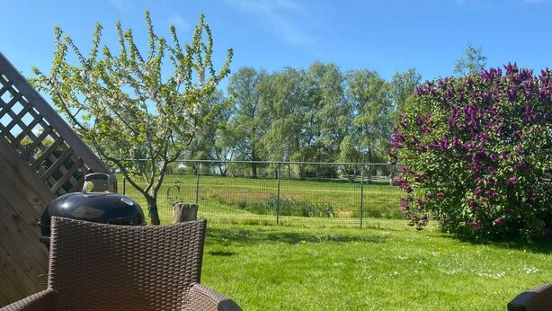 Een achtertuin met een barbecue, rieten stoelen, een houten terras, een omheining en een tuin met bomen en bloemen.