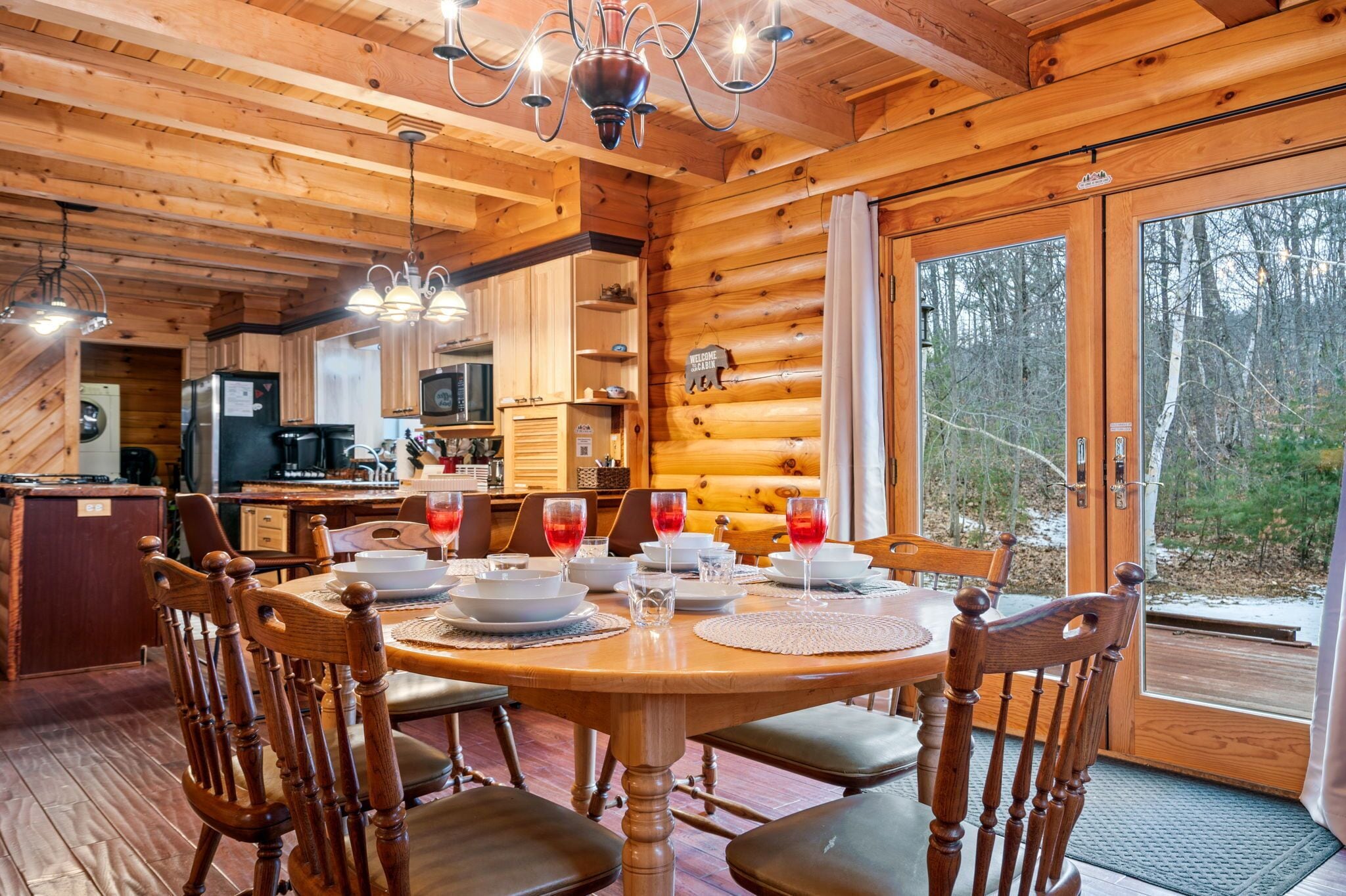A wooden cabin interior with a dining table set for four, a kitchen area, and a large window offering a view of the forest.