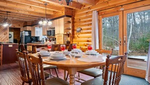 A wooden cabin interior with a dining table set for four, a kitchen area, and a large window offering a view of the forest.