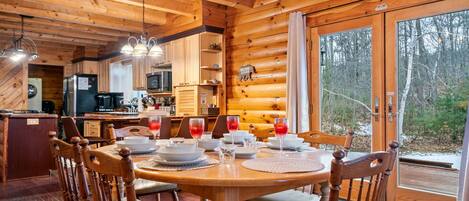 A wooden cabin interior with a dining table set for four, a kitchen area, and a large window offering a view of the forest.