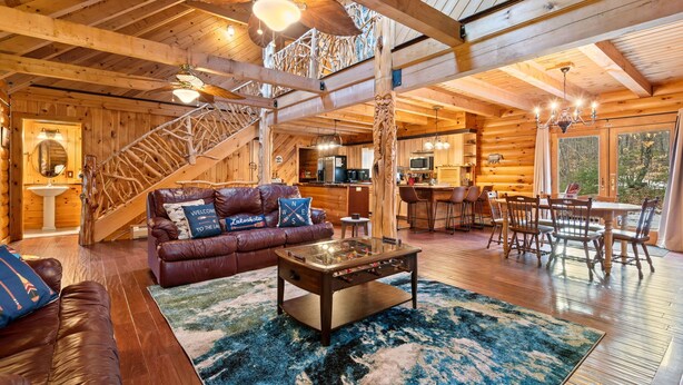 A log cabin interior with a wooden staircase, a living room with a sofa and coffee table, a dining area with chairs, and a kitchen in the background.