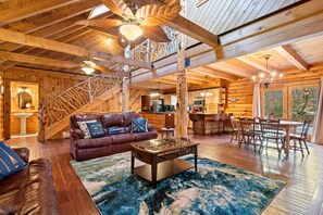 A log cabin interior with a wooden staircase, a living room with a sofa and coffee table, a dining area with chairs, and a kitchen in the background. - Warm Lakefront Log Cabin · Hot Tub · Game Room (Wakefield)