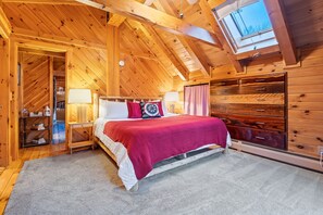 Ein gemütliches Schlafzimmer mit Holzbalkendecke, einem Bett mit roter Decke, einem Nachttisch und einem Fenster mit Blick zum Himmel.