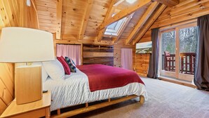 A wooden cabin bedroom with a bed, nightstand, lamp, and a view of the outdoors through a large window.