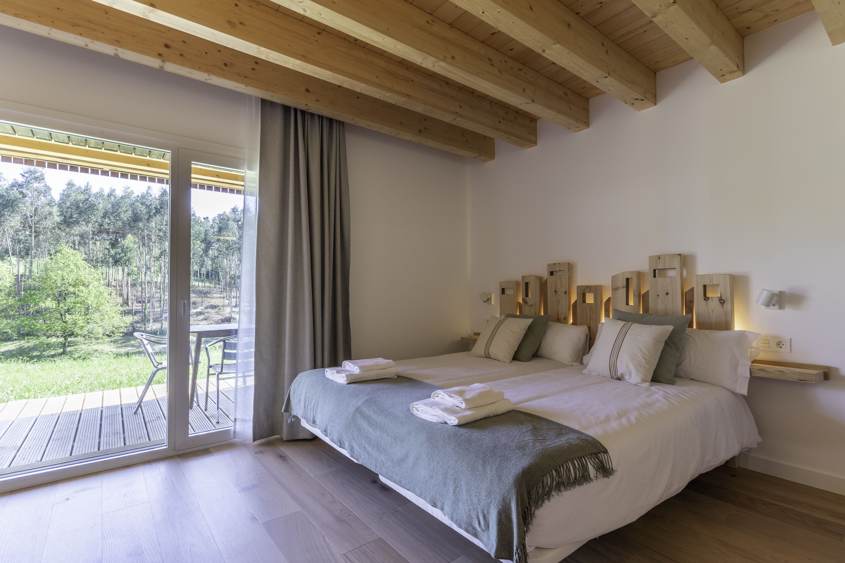 Un dormitorio con una cama grande, techo de madera, puerta de vidrio corredera a un balcón y vistas a árboles.