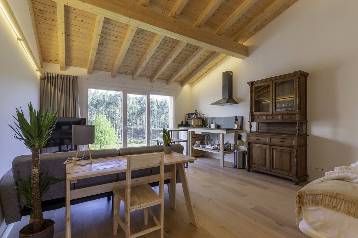 Uma sala de estar ampla com teto de madeira, área de jantar, cozinha e uma grande janela com vista para o exterior.