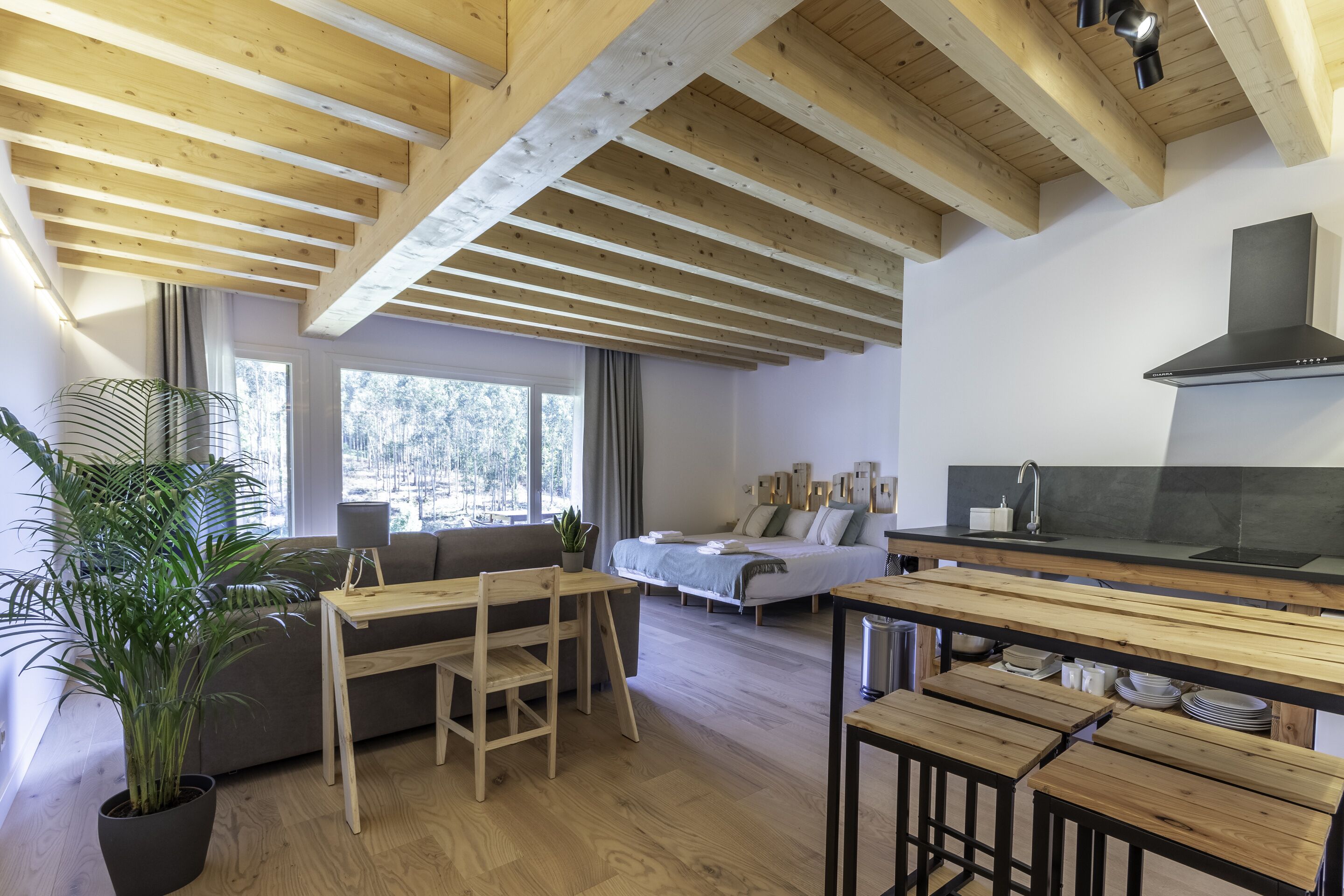 A modern, open-plan living space with a wooden ceiling, a dining area with benches, a sofa, a bed, and a kitchen area with a black stove.