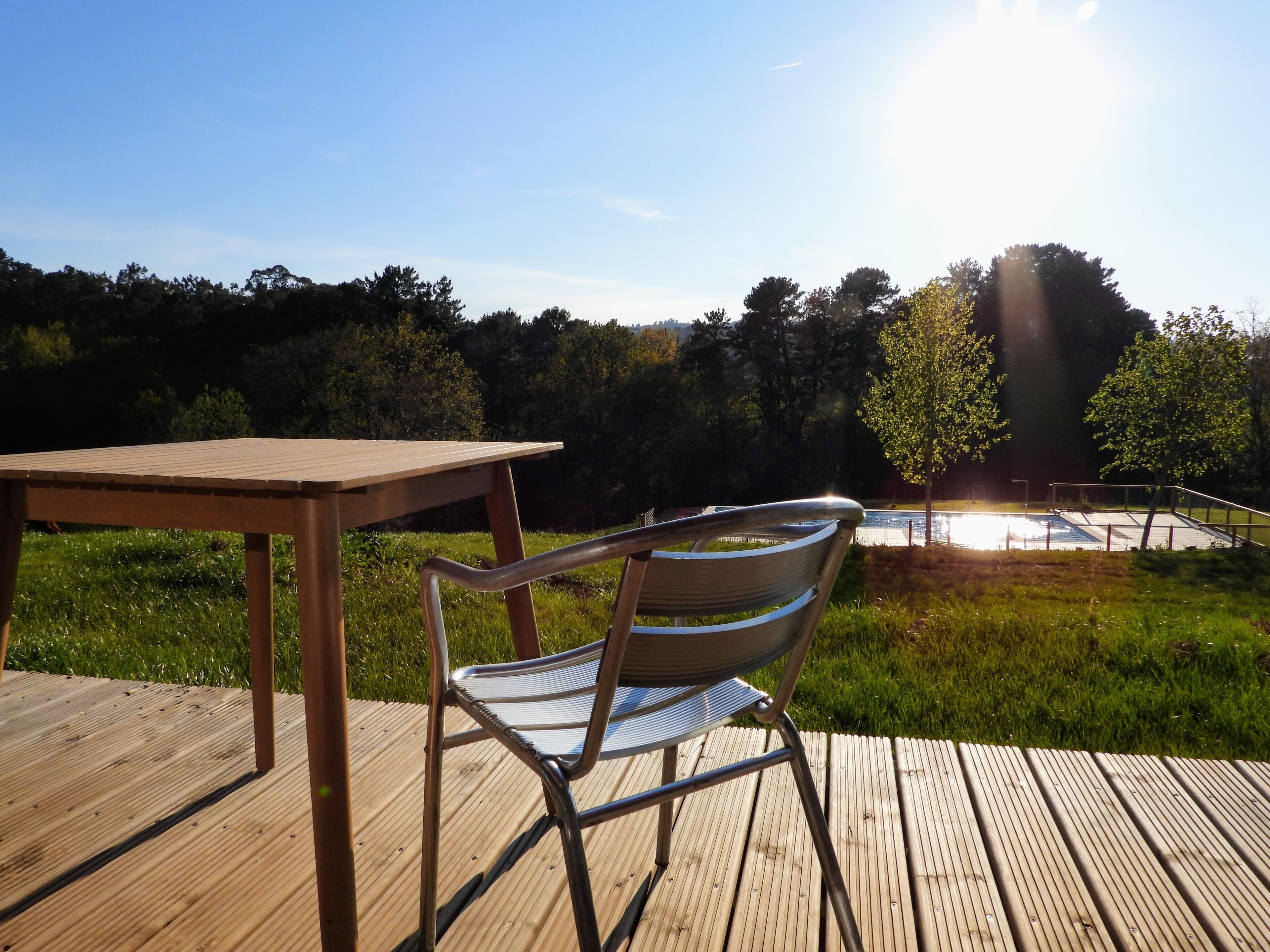 A wooden deck with a metal chair and a table, overlooking a swimming pool and trees.