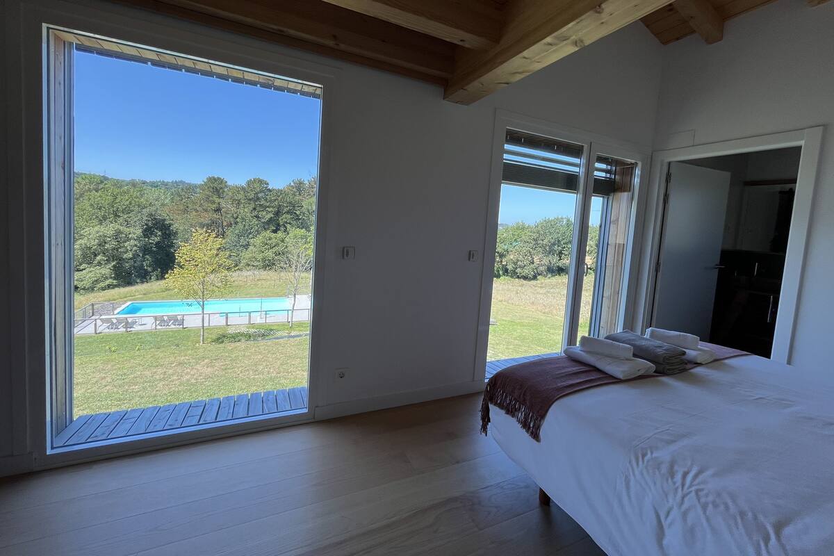 Quarto com uma grande porta de vidro deslizante que dá para uma varanda, cama com edredom branco e vista para uma piscina e vegetação.